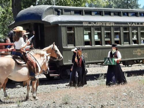 A group of people in Western attire near a vintage train, with one person on a horse and others holding props.