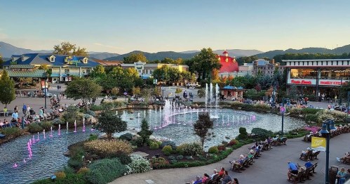A scenic park with a pond, colorful fountains, and people enjoying the outdoors, surrounded by mountains and buildings.