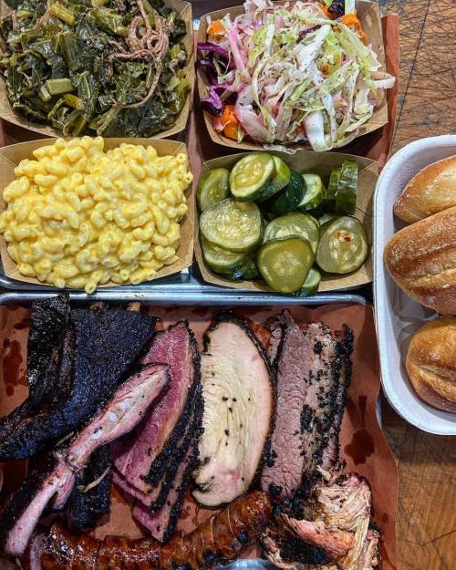 A tray of barbecue featuring brisket, sausage, pulled pork, sides of macaroni, pickles, greens, and coleslaw, with rolls.