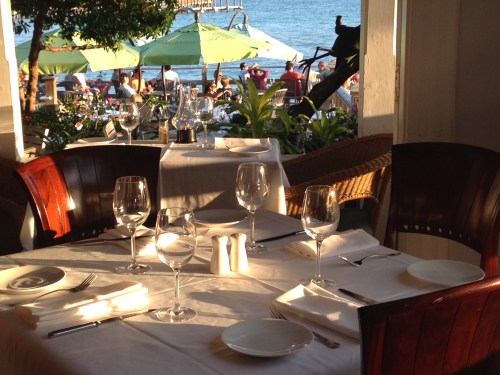 A beautifully set dining table with wine glasses, overlooking a beach scene with umbrellas and people in the background.