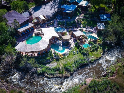 Aerial view of a resort with pools and cabanas beside a flowing river, surrounded by lush greenery.