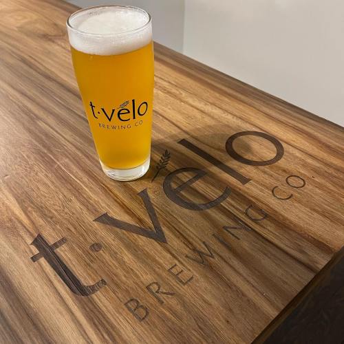 A glass of golden beer sits on a wooden table with the logo of t. velo Brewing Co. visible.