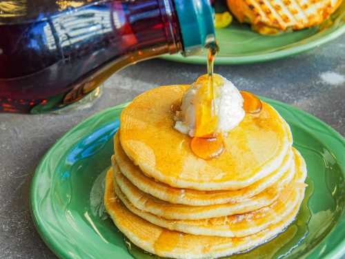 A stack of fluffy pancakes topped with a scoop of butter and drizzled with syrup on a green plate.