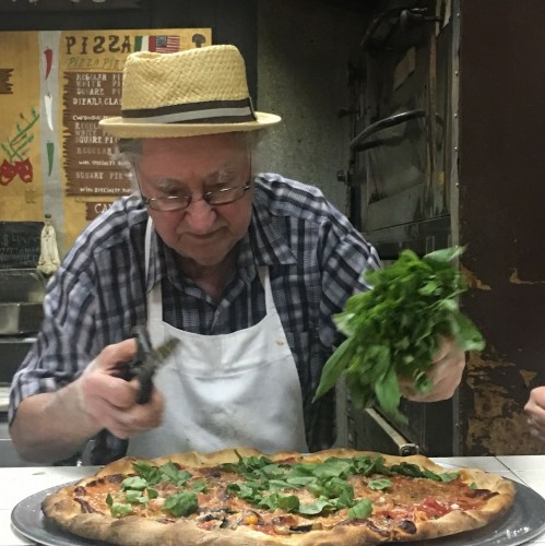An elderly man in a straw hat and apron prepares a pizza, adding fresh greens in a rustic kitchen.