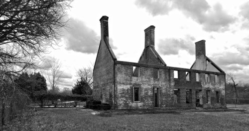 Black and white image of a partially collapsed brick house with two tall chimneys, surrounded by bare trees and cloudy skies.
