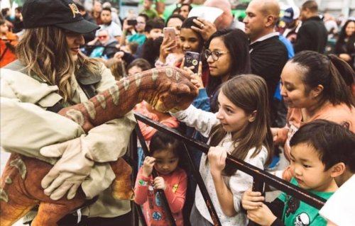 A crowd of excited children and adults watch as a person holds a dinosaur puppet at an event.