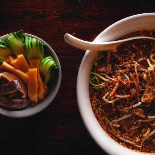A bowl of spicy noodles topped with vegetables and sesame seeds, alongside a small dish of sliced cucumbers and mushrooms.