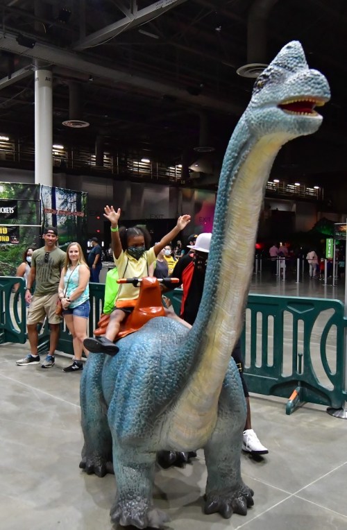 A child joyfully rides a large dinosaur statue, while a group of people smiles in the background at an indoor event.