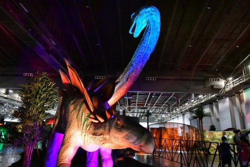 A large dinosaur model with a long neck, illuminated in colorful lights, set in a dimly lit exhibition space.