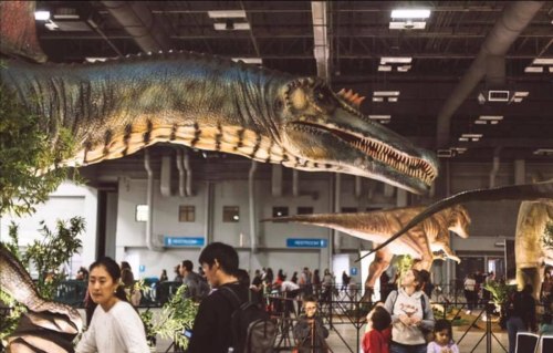 A dinosaur exhibit featuring large dinosaur models with visitors exploring the display in a spacious indoor venue.