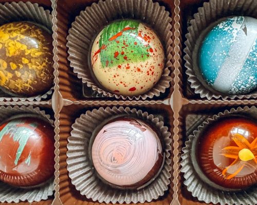 A box of colorful, artistic chocolates arranged in a grid, featuring various vibrant designs and patterns.