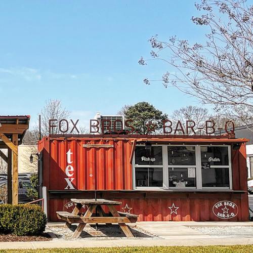 A red shipping container restaurant with a sign reading "Fox Bros. Bar-B-Q," featuring a picnic table outside.