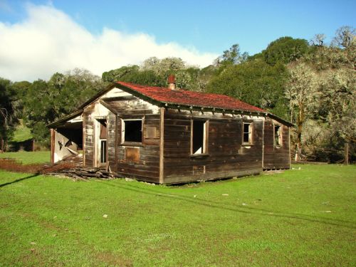 Abandoned wooden house with a red roof, missing windows, surrounded by green grass and trees under a blue sky.