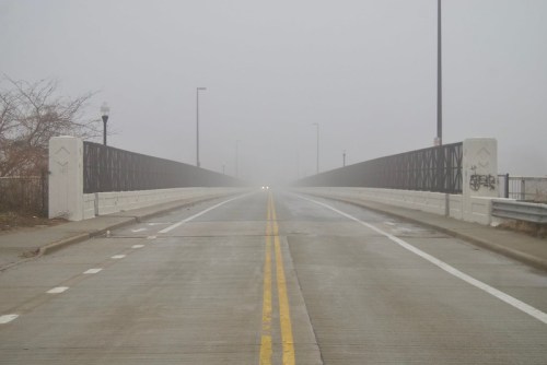A foggy view of a bridge with a two-lane road, disappearing into the mist ahead. Streetlights are faintly visible.