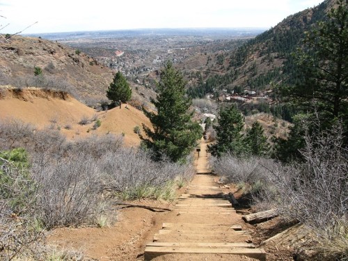 A steep, wooden staircase leads down a hillside, surrounded by trees and shrubs, with a valley visible in the distance.
