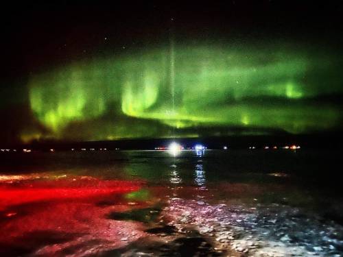 A stunning display of green auroras dances over a dark lake, reflecting lights in the water below.