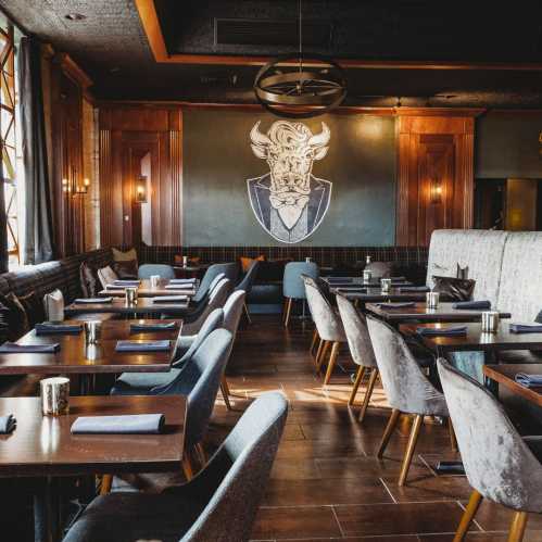 Interior of a stylish restaurant featuring wooden tables, upholstered chairs, and a large bull mural on the wall.