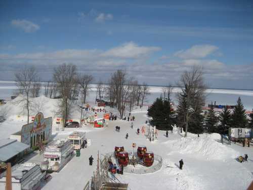 A snowy landscape with amusement rides, food stalls, and people enjoying a winter festival by a frozen lake.