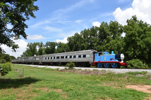 A blue and red train resembling a cartoon character travels along a track beside green trees and a grassy area.