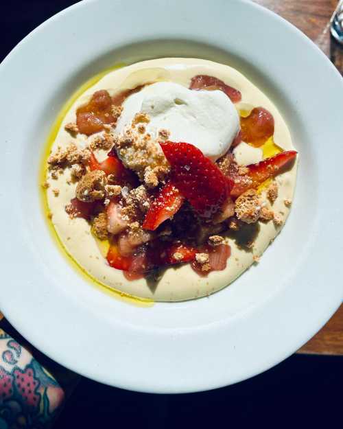 A creamy dessert topped with strawberries, rhubarb, crumbled cookies, and a dollop of whipped cream in a white bowl.