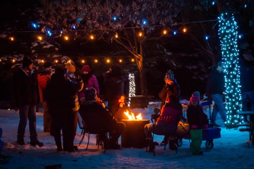A group of people gathered around a fire in a snowy setting, with colorful lights strung in the background.