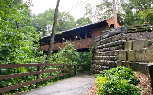 A wooden covered bridge spans over a lush, green path surrounded by trees and stone walls.