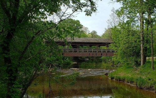 A covered bridge spans a serene creek, surrounded by lush green trees and a peaceful natural landscape.