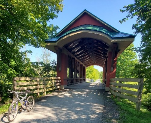 A covered bridge surrounded by greenery, with a bicycle parked on the side of a path leading through it.