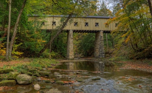 A wooden covered bridge spans a creek, surrounded by trees with autumn foliage. Rocks line the water's edge.