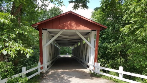 A red-roofed covered bridge surrounded by lush green trees and a white fence, with a path leading through it.