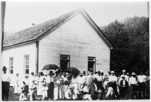 A large group of people, some with umbrellas, gathered in front of a small wooden building in a rural setting.