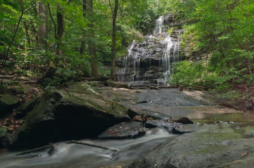 A serene waterfall cascades over rocks, surrounded by lush green trees and a calm stream in a tranquil forest setting.