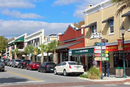 A charming street lined with shops and palm trees, featuring parked cars and a clear blue sky.