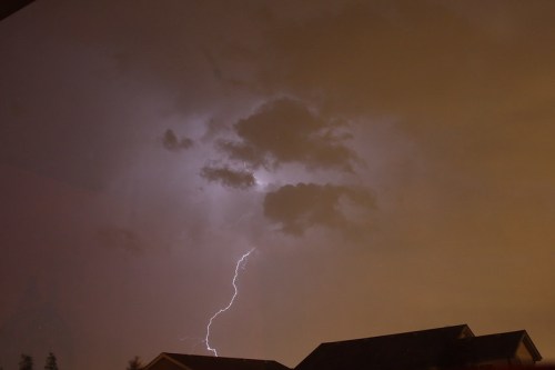 A dramatic sky with dark clouds illuminated by lightning, silhouetting rooftops below.