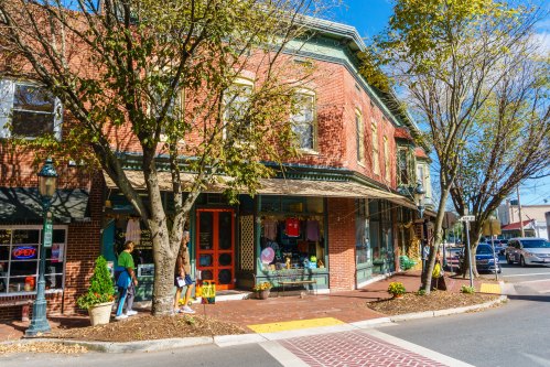 A charming brick storefront with trees, colorful displays, and an "Open" sign on a sunny day in a small town.