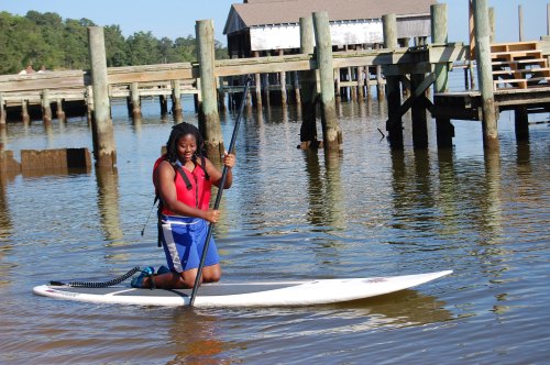A person kneels on a paddleboard in calm water near a dock, wearing a life jacket and smiling.