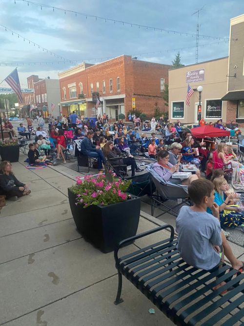 A large crowd sits on benches and blankets outdoors, enjoying an event in a small town, with flowers and buildings in view.