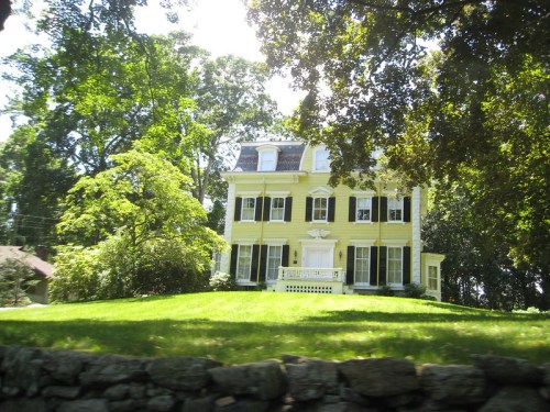 A large yellow house with white trim, surrounded by lush green trees and a manicured lawn.