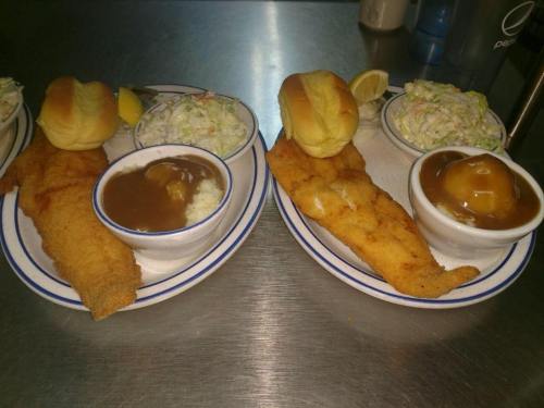 Two plates of fried fish with sides of coleslaw, rice, and rolls, garnished with lemon wedges.
