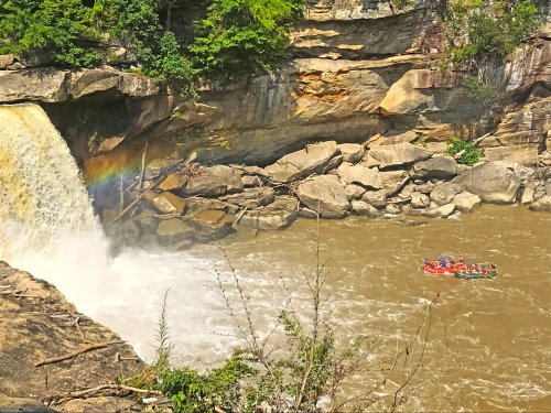 A group of people rafting near a waterfall, with a rainbow visible in the mist and rocky cliffs surrounding the scene.