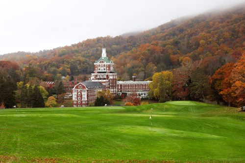 A grand hotel surrounded by colorful autumn foliage and a green golf course, set against a misty mountain backdrop.