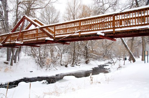 A wooden bridge covered in snow spans a frozen creek, surrounded by snow-laden trees in a winter landscape.