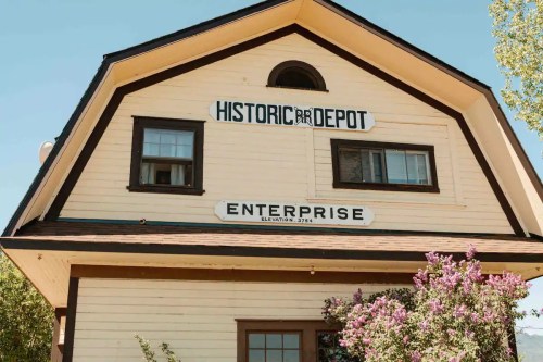 Historic depot building with "Enterprise" sign, featuring a peaked roof and blooming flowers in front.