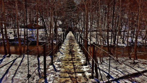 A snowy landscape featuring a wooden suspension bridge over a stream, surrounded by bare trees.