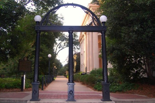 A decorative archway with lights, leading to a pathway surrounded by trees and a historic building in the background.