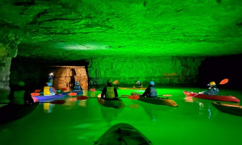 A group of kayakers paddles in a glowing green cave, illuminated by vibrant lights reflecting off the water.