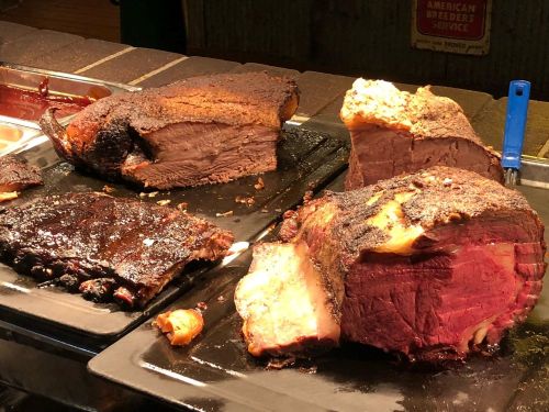 A selection of smoked meats on black trays, including brisket and ribs, with a charred, flavorful crust.