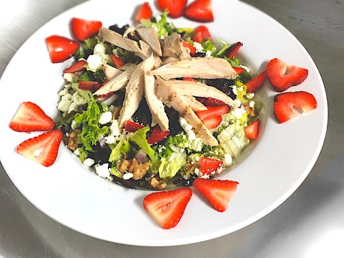A colorful salad with mixed greens, sliced chicken, strawberries, and crumbled cheese, served in a white bowl.
