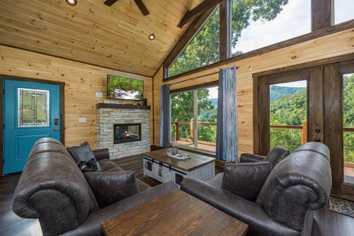 Cozy living room with wooden walls, a stone fireplace, large windows, and a view of mountains.