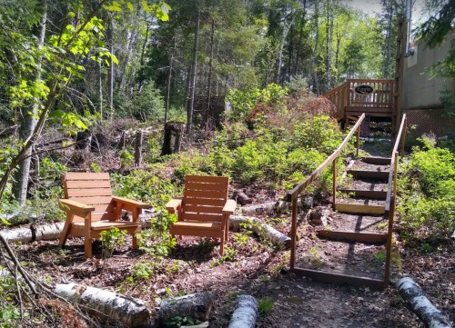 Two wooden chairs sit in a forested area, with a staircase leading up to a deck in the background.